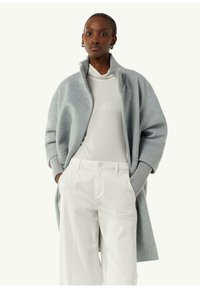 Grauer Wollmantel mit hohem Kragen und übergroßen Ärmeln, kombiniert mit einem weißen Rollkragenpullover und weit geschnittenen weißen Hosen. Glatte Textur.