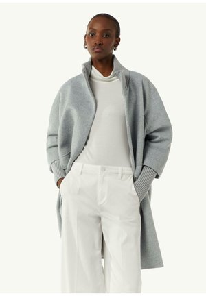 Grauer Wollmantel mit hohem Kragen und übergroßen Ärmeln, kombiniert mit einem weißen Rollkragenpullover und weit geschnittenen weißen Hosen. Glatte Textur.