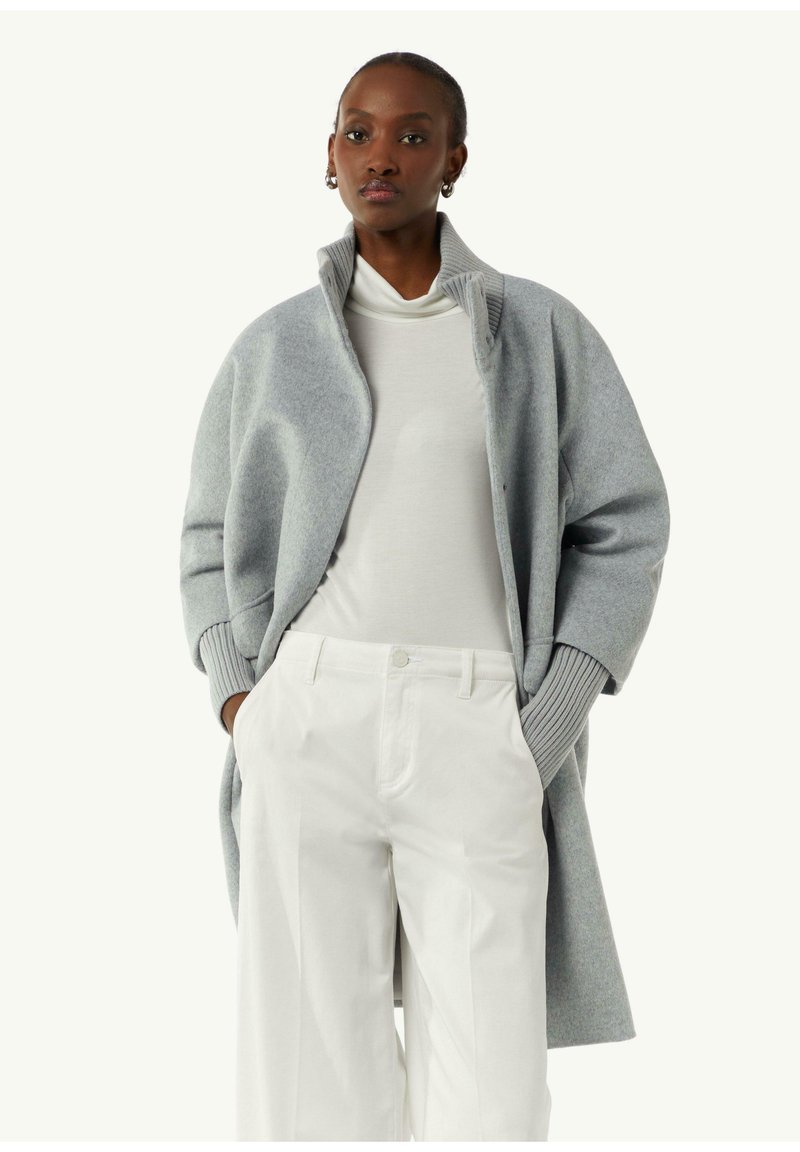 Grauer Wollmantel mit hohem Kragen und übergroßen Ärmeln, kombiniert mit einem weißen Rollkragenpullover und weit geschnittenen weißen Hosen. Glatte Textur.