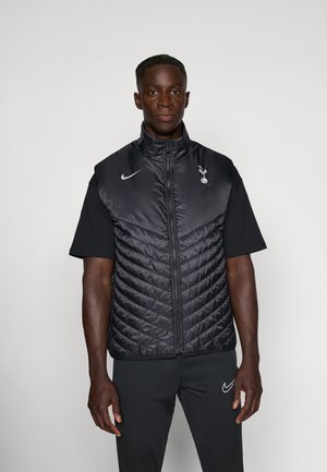 Black puffer vest with a high collar, featuring diagonal quilted pattern and a Nike logo. Worn over a black short-sleeve shirt.