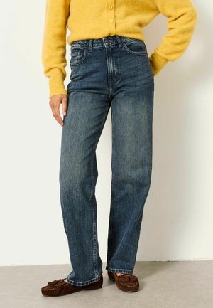 Person, die blaue Jeans mit hoher Taille und geradem Bein, braune Mokassins und einen gelben, flauschigen, knöpfbaren Pullover trägt, steht vor einer weißen Wand.