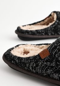 Chaussons tricotés noirs et gris avec une doublure en peluche beige et une étiquette en cuir marron. Forme arrondie avec un extérieur texturé. Semelle plate.