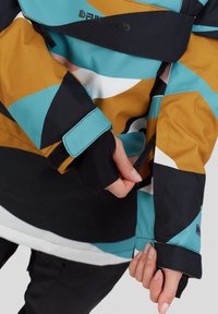Kolorowa kurtka z wyrazistymi geometrycznymi wzorami w kolorach morskim, musztardowym, czarnym i białym, z regulowanymi mankietami i eleganckim fasonem.