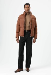 Bruine pufferjas met een hoge kraag, gecombineerd met een beige coltrui en zwarte wijde jeans, gedragen met bruine leren schoenen.