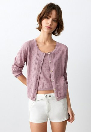 Giovane donna che indossa un cardigan lavorato a maglia color lavanda e un top corto abbinato con pantaloncini bianchi a vita alta, in piedi su uno sfondo bianco uniforme.