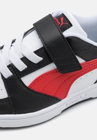 Basket noire et blanche avec des accents rouges, en matériau en cuir texturé, présentant une sangle en Velcro et un design de lacets classique.