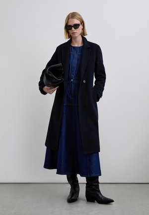 Abrigo negro de lana con solapas de muesca, combinado con un vestido de mezclilla. La modelo sostiene un bolso de cuero negro, lleva gafas de sol negras angulares y botas hasta la rodilla.