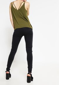 Nainen pukeutuneena oliivinvihreään hihattomaan toppiin, jossa on ohuet olkaimet, mustiin kapeisiin farkkuihin ja mustiin korotettuihin korkokenkiin, seisoo selkä kääntyneenä.