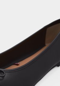 Ballerina in pelle nera con punta arrotondata, piccolo fiocco decorativo e fodera interna color beige su sfondo bianco.