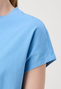 Detailný záber na osobu v svetlo modrom tričku s krátkym rukávom z bavlny, ukazujúcim rameno a hornú časť ramena na jednoduchom pozadí.
