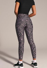 Svarta leggings med ett virvlande lila mönster, hög midja och elastiskt material, tillsammans med svarta sportskor.
