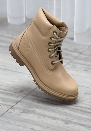 Bottine en cuir tan avec bout rond, lacets beige, œillets en métal et semelle en caoutchouc. Présente un logo embossé et un panneau latéral texturé.
