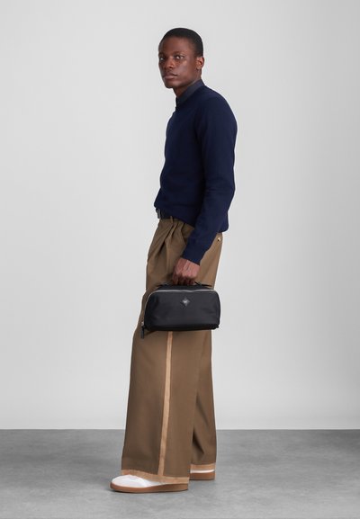 Czarna, zapinana torba o prostokątnym kształcie, z akcentem w postaci logo. Noszona z granatowym swetrem i beżowymi szerokimi spodniami, w zestawieniu z białymi butami.