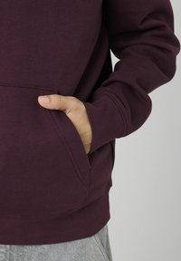 Sweatshirt bordeaux avec une poche avant, des poignets côtelés et une texture douce. Le tissu semble épais et chaud, idéal pour un port décontracté.