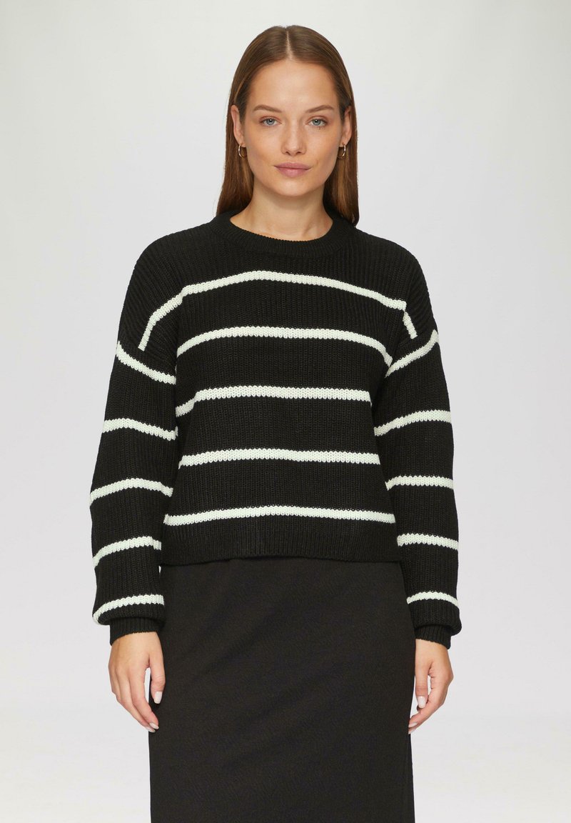 Schwarzer Strickpullover mit horizontalen weißen Streifen, rundem Ausschnitt und langen Ärmeln. Kombiniert mit einem schwarzen strukturierten Rock.