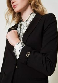 Zwarte op maat gemaakte blazer met een gestructureerde pasvorm, gecombineerd met een witte blouse met een zwart dierenprint. Gouden hardware accenten op de manchetten.