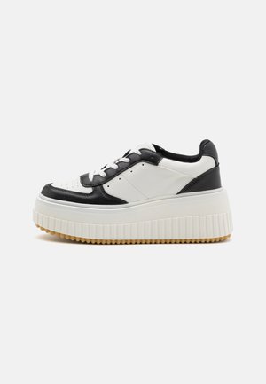 Sneaker plateforme noir et blanc avec une semelle épaisse à nervures et des lacets blancs, présenté de profil sur un fond clair.