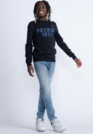Marineblauwe trui met "PETROL 1973" in blauwe letters, gecombineerd met lichtblauwe jeans en witte sneakers. De stof lijkt zacht met geribde boorden.