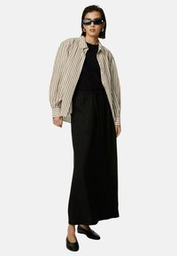 Gestreepte beige en bruine oversized shirt, zwarte top en zwarte maxi rok; zwarte platte schoenen; model draagt zwarte zonnebril en oorbellen.