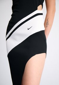 Svart og hvit atletisk kjole med utskjæringsdetalj, preget av et diagonalt stripete design, laget av elastisk stoff, inkluderer en liten Nike-logo.