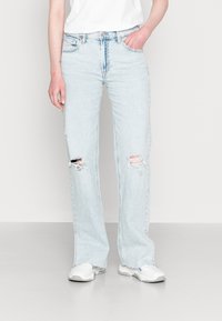 Jeans azul claro, de corte solto, com rasgos nos joelhos, cinco bolsos, passantes para cinto e bainhas desfiadas; combinados com ténis brancos.