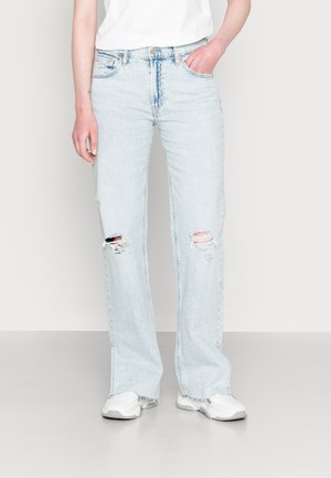 Jeans azul claro, de corte solto, com rasgos nos joelhos, cinco bolsos, passantes para cinto e bainhas desfiadas; combinados com ténis brancos.
