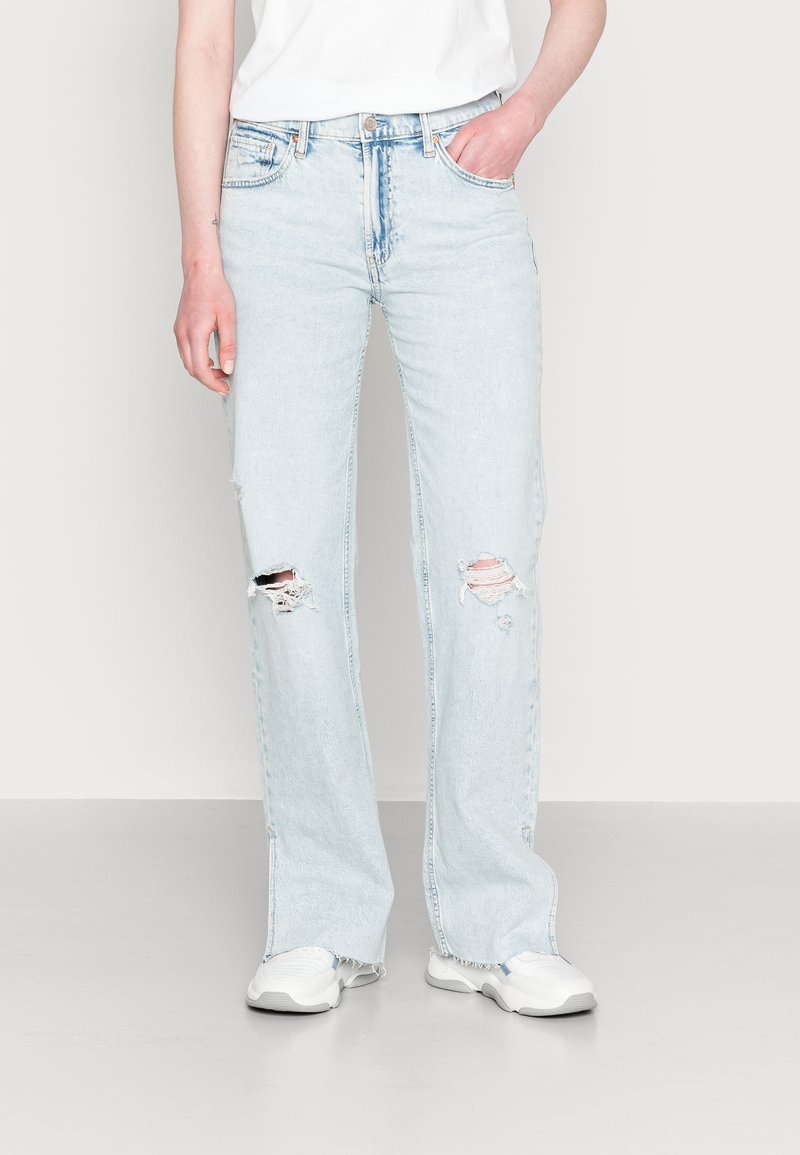 Jeans azul claro, de corte solto, com rasgos nos joelhos, cinco bolsos, passantes para cinto e bainhas desfiadas; combinados com ténis brancos.
