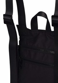 Bolsa de tela negra con dos bolsillos con cremallera, correas negras ajustables y un asa de transporte en la parte superior sobre un fondo blanco.