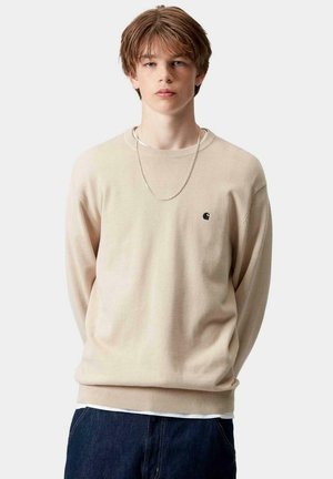 Joven con cabello castaño claro que lleva un suéter beige con un pequeño logo, cadena de plata y jeans azul oscuro, de pie contra un fondo liso.