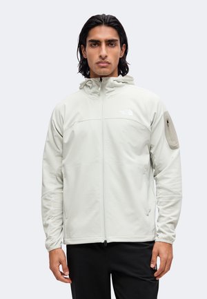 Homme aux cheveux noirs mi-longs portant une veste gris clair zippée avec une capuche et une poche zippée sur la manche gauche.