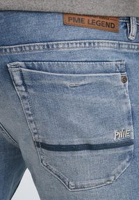 Světle modré džíny z denimu s vybledlou texturou, s hnědou koženou etiketou označenou "PME LEGEND" a kontrastním tmavě modrým pruhem na zadní kapse.