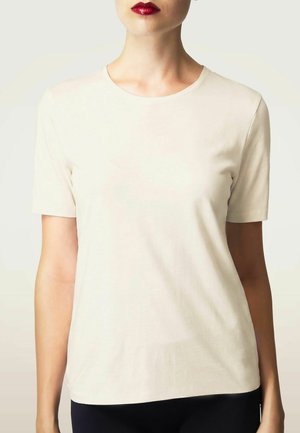 Vrouw met rode lippenstift die een effen lichtbeige korte mouw T-shirt en donkere broek draagt, bij schouders en taille bijgesneden.