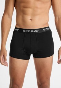 Boxer infradito neri con un morbido elastico in vita che presenta il logo "GUESS JEANS". Il materiale è liscio, aderente e leggero.