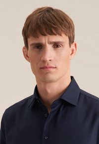 Camicia blu navy con bottoni, colletto a punta, texture liscia e motivo sottile, indossata da un modello maschile su uno sfondo neutro.
