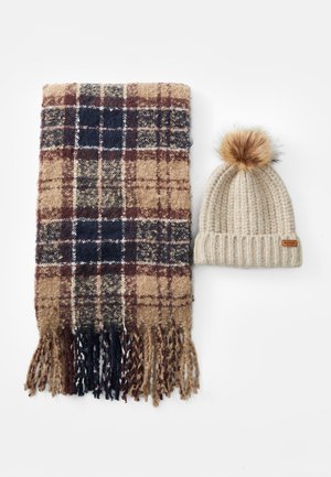 Beige en marineblauw geruit sjaal met franjes, gecombineerd met een lichtbeige ribgebreide beanie met een bruin leren label en een fluffy pompon.