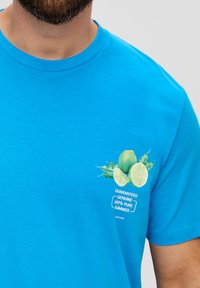 Turquoise katoenen t-shirt met korte mouwen, met een afbeelding van limoenen en de tekst "GEGARANDEERD ECHT 100% PURE ZOMER."