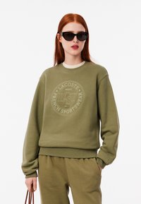 Sudadera verde oliva con un logotipo texturizado, puños acanalados y cuello redondo, combinada con pantalones a juego y gafas de sol negras.