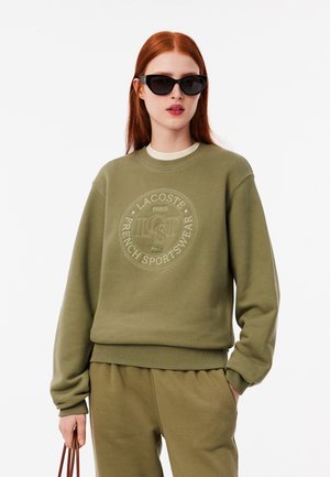 Sudadera verde oliva con un logotipo texturizado, puños acanalados y cuello redondo, combinada con pantalones a juego y gafas de sol negras.
