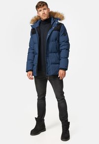 Marineblå puffjakke med pelsforet hette, sorte detaljer på skuldrene, glidelåslukking og quiltet design. Modell som har på seg svarte jeans og støvler.