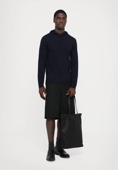 Felpa con cappuccio navy, pantaloni corti neri, scarpe di pelle nere e una borsa a tracolla nera. La felpa presenta una tasca frontale e polsini a coste.