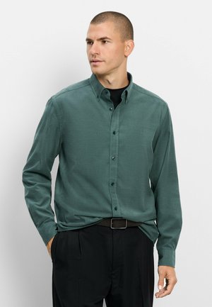 Mann trägt dunkelgrünes Hemd mit Knöpfen über schwarzem Top, schwarze Hose und Gürtel, steht mit einer Hand in der Tasche und blickt nach links.