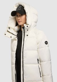 Biela vatovaná bunda s kapucňou, so zapínaním na zips vpredu, čiernymi detailmi a veľkými prešívanými časťami. Nosená s čiernou šiltovkou.