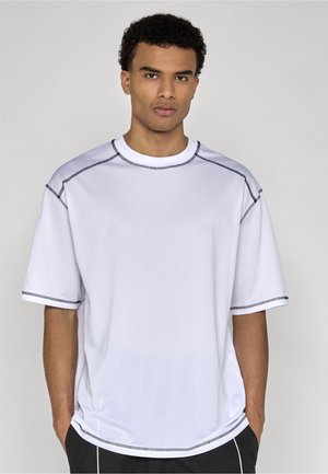 Jeune homme aux cheveux courts et bouclés portant un T-shirt blanc ample avec des coutures noires, debout les mains dans les poches devant un fond uni.