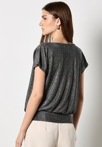 Schwarzes Glitzeroberteil mit kurzen Ärmeln und lockerer Passform, das mit drapiertem Stoff und einem enganliegenden Taillenbund begeistert. Das Material hat eine schimmernde Struktur.