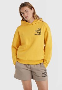 Żółta bluza z kapturem z czarnym napisem "The Future Surf Society" z przodu, w połączeniu z beżowymi szortami z tym samym czarnym napisem. Miękki materiał.