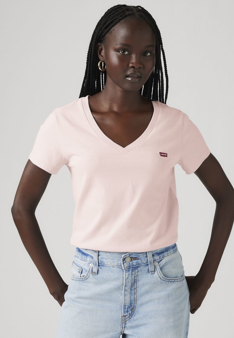 Femme aux cheveux tressés portant des boucles d'oreilles créoles dorées, un t-shirt rose clair à col en V avec un logo rouge, et un jean taille haute bleu clair.
