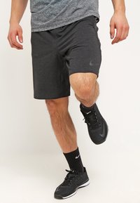 Hombre vistiendo pantalones cortos atléticos gris oscuro, calcetines negros de Nike y zapatillas negras de Nike, equilibrándose en un pie contra un fondo sencillo.