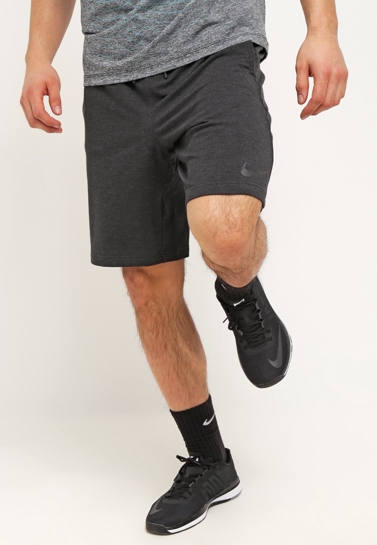 Hombre vistiendo pantalones cortos atléticos gris oscuro, calcetines negros de Nike y zapatillas negras de Nike, equilibrándose en un pie contra un fondo sencillo.