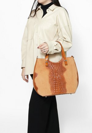 Borsa a tracolla in pelle marrone chiaro con motivo rettile testurizzato, finitura liscia e manici robusti; indossata con una camicia crema e pantaloni neri.