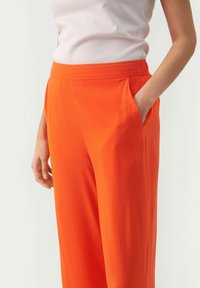 Orange vide benbukser med en glat tekstur, der har sidelommer og en elastisk talje. Kombinationen er med en lyserød top.